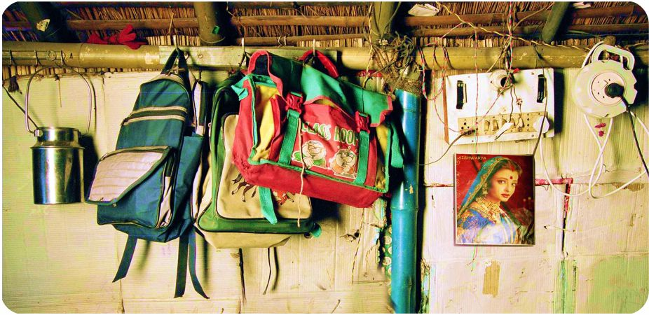 दीवार पर स्कूल के बैग, बर्तन, बिजली की तार, पोस्टर लगा है।