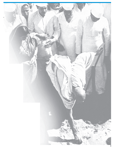 डांडी यात्रा की तस्वीर जिसमे गांधी जी तट से मुट्ठी में नमक भर रहे हैं और कई लोग उनके पास खड़े हैं।