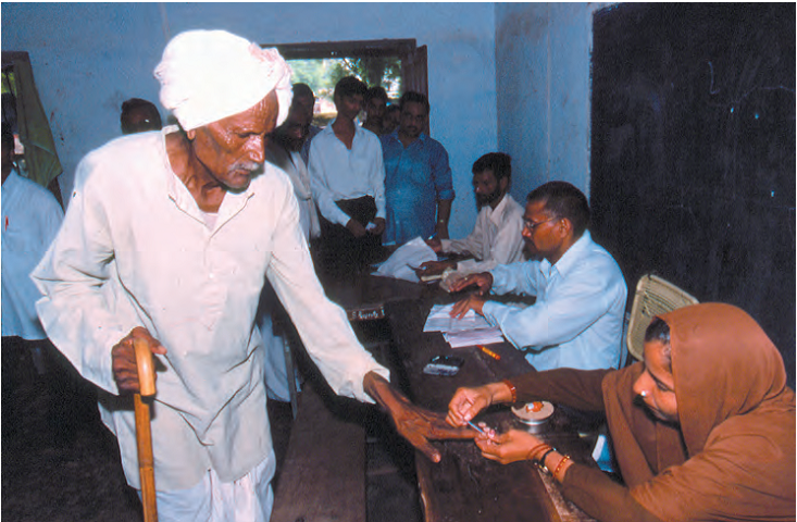 मतदान कक्ष में वृद्ध की अंगुली पर निशान लगाती महिला। वार्ड में मतदाताओं की लंबी कतार है।