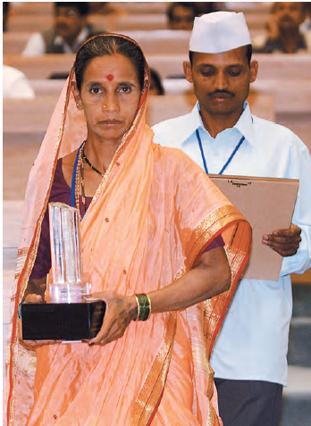 एक महिला अपने हाथ में पुरस्कार लिए हुए है, जबकि एक आदमी अपने हाथ में लकड़ी का बोर्ड लिए हुए है, उसके पीछे खड़ा है।