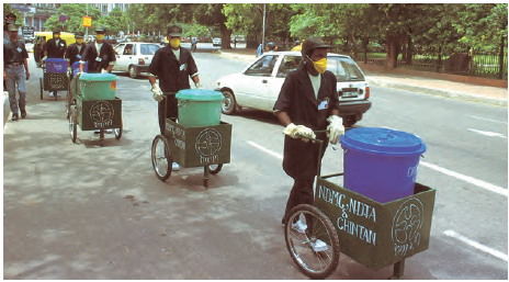   एक सड़क के किनारे, कुछ आदमी छोटी गाड़ियों पर बड़े कूड़ेदान के बंद  बक्से ले जा रहे हैं। पुरुष मास्क पहने हुए हैं और उनके हाथ दस्ताने से ढके हुए हैं।  