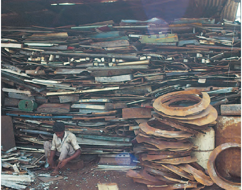 एक जगह के फर्श पर बैठा एक आदमी, जहाँ ढेर सारा प्लास्टिक, धातु, कागज  और कांच रखे हुए हैं।  