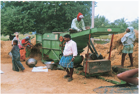 कुछ लोग खेत में काम कर रहे हैं और खेत में ट्रैक्टर जैसी बड़ी मशीन भी है और उसके ऊपर एक आदमी बैठा है।