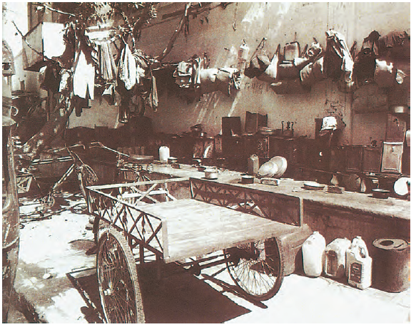 गली के एक किनारे पर एक खाली ठेला खड़ा है और पास ही एक छोटी सी रसोई बनी है और वहीं उपर तारों पर कुछ कपड़े भी टंगे हैं। 