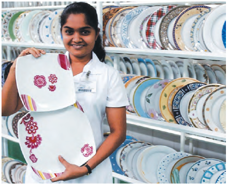 एक दुकान में बेचने के लिए एक लड़की दो प्लेट दिखा रही है। उसके पीछे रैक में कई प्लेट रखी हुई हैं।  