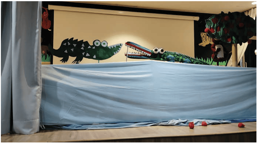 A bedsheet covering a table is used as a stage. The bedsheet will hide the puppeteer, who can move the puppets using their hands or sticks attached to the puppets’ bodies.