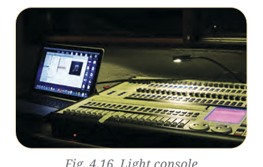 A high-end digital lighting console illuminated in a dark theater booth next to a laptop control screen.